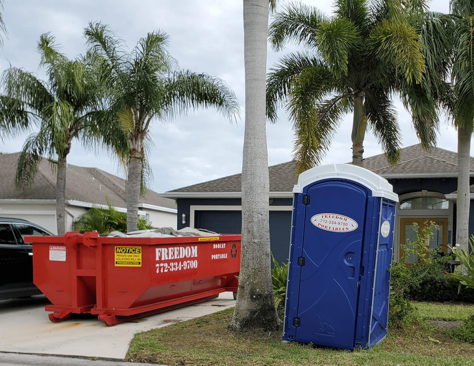 a red dumpster in a driveway and a blue porta potty in a front yard